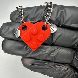 May include: A red heart-shaped pendant made from two interlocking brick pieces. Each piece has a silver chain attached. One piece has the letter "M" and the other has the letter "U" on a small silver charm. The pendant is a friendship or couple's necklace.