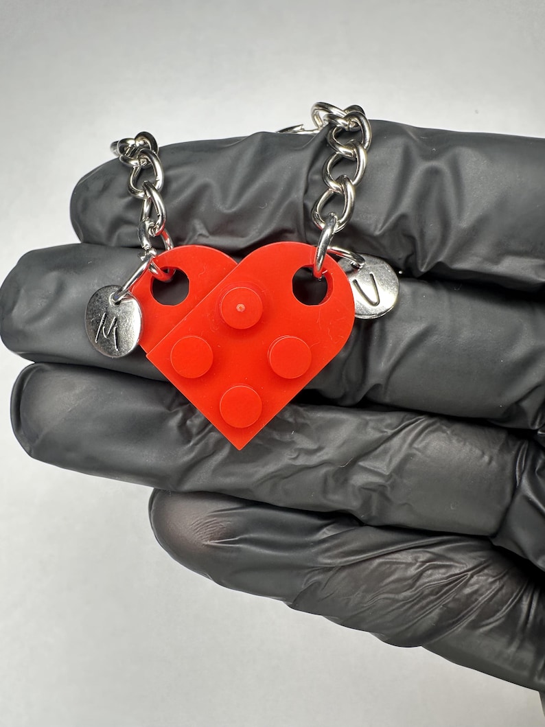 May include: A red heart-shaped pendant made from two interlocking brick pieces. Each piece has a silver chain attached. One piece has the letter "M" and the other has the letter "U" on a small silver charm. The pendant is a friendship or couple's necklace.