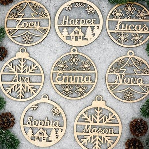 May include: Wooden Christmas ornaments with personalized names, snowflakes, and tree designs. Each ornament is circular with a loop for hanging. Names include Zoey, Harper, Lucas, Ava, Emma, Nova, Sophia, and Mason. The ornaments are surrounded by pine needles and pine cones.