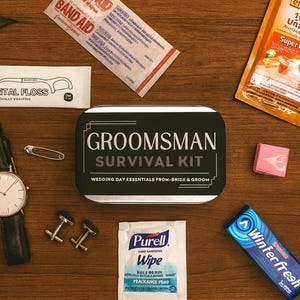 May include: A flat lay of a groomsman survival kit with various wedding day essentials. The kit includes a watch, cufflinks, dental floss, band-aids, hand sanitizer wipes, and Emergen-C packets. The kit is in a black tin with the text "GROOMSMAN SURVIVAL KIT".