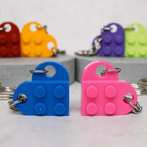 May include: A selection of heart-shaped keyrings in various colours, including red, yellow, purple, green, blue, pink, and grey. Each keyring is designed to look like a building block, with a metal ring for keys. A fun and unique accessory.