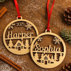 May include: Two wooden Christmas ornaments with red ribbons. Each ornament is round with a cut-out design featuring a house, trees, snowflakes, and a name. One says "Harper" and the other says "Sophia". The year "2025" is also visible.