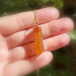 May include: A gold chain necklace with a long, orange carnelian pendant. The pendant is faceted and has a pointed tip.