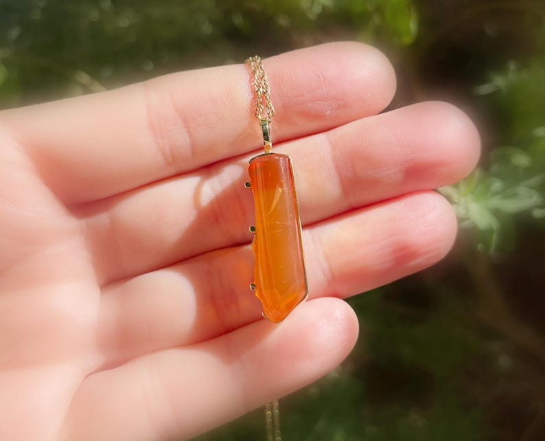 May include: A gold chain necklace with a long, orange carnelian pendant. The pendant is faceted and has a pointed tip.