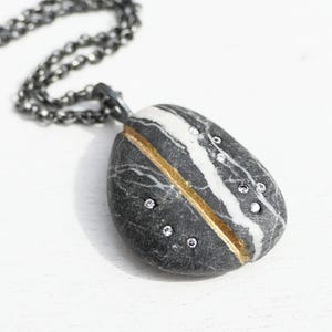 May include: A pendant necklace featuring a polished, oval-shaped stone with a black, white, and gold design. The stone is adorned with small, sparkling crystals. The necklace has a dark, chain.