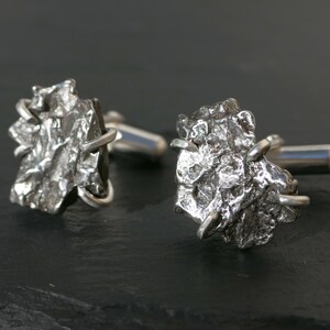 May include: A pair of silver cufflinks with a unique, textured design. The cufflinks have a rugged, organic appearance, with a metallic sheen. The post and backing are smooth and polished. The cufflinks are displayed on a dark surface.