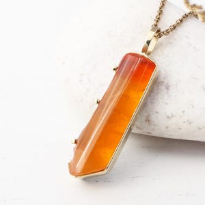 May include: A gold chain necklace with a pendant featuring a long, orange, faceted gemstone.