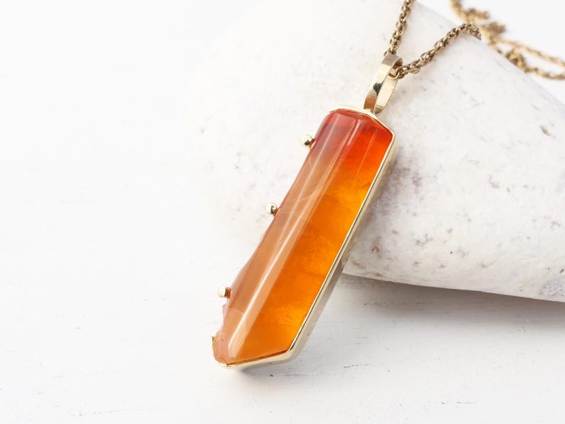 May include: A gold chain necklace with a pendant featuring a long, orange, faceted gemstone.