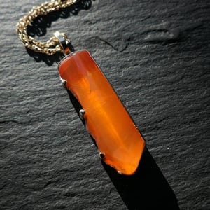 May include: A gold chain necklace with a pendant featuring a long, orange carnelian gemstone.