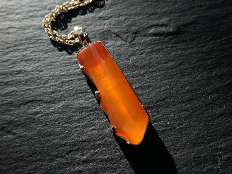May include: A gold chain necklace with a pendant featuring a long, orange carnelian gemstone.