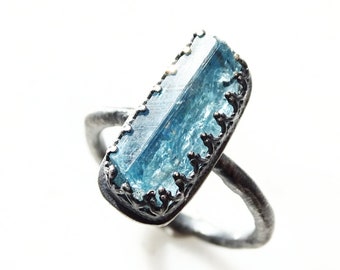 May include: A silver ring featuring a rectangular, light blue gemstone. The gemstone is set in a decorative, dark silver bezel. The ring has a textured band and a unique, artistic design. The ring is a piece of jewelry.