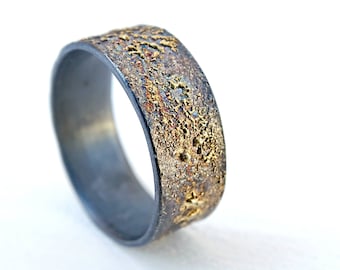 May include: A black and gold textured metal ring. The ring has a wide band with a rough, uneven surface.