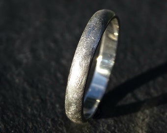 May include: A silver ring with a textured, brushed finish. The ring has a smooth, rounded band and reflects light, creating highlights. The ring is a simple, elegant piece of jewelry.
