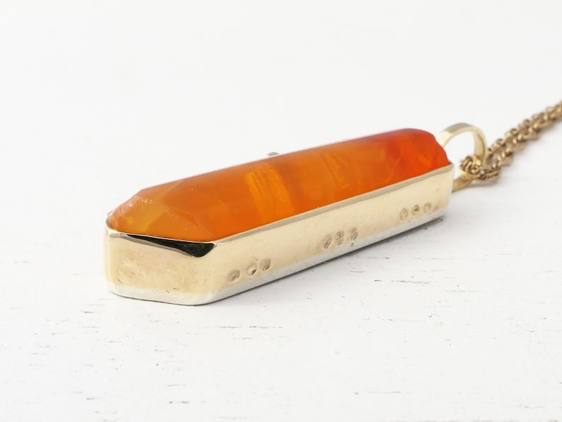 May include: A gold pendant necklace with an orange crystal shaped like a long, thin point. The pendant is set in a gold frame and has a detailed design.