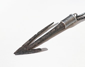 May include: A rustic, metal spearhead pendant necklace. The pendant features a dark, weathered spearhead design with a silver-colored metal accent and chain. The spearhead has a three-pronged design.