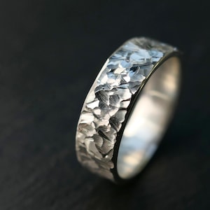 May include: A silver ring with a textured, hammered finish. The ring has a wide band and a shiny, reflective surface. The ring is set against a dark background, highlighting its metallic sheen. A piece of jewelry.