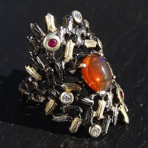 May include: A silver ring with a large orange opal gemstone set in a black and gold textured band. The band is decorated with smaller red and white gemstones.