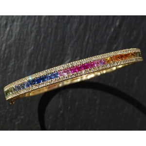 May include: A gold bangle bracelet with a rainbow row of square-shaped gemstones surrounded by a row of smaller round gemstones.