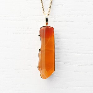May include: A gold chain necklace with a pendant made of a large, orange carnelian gemstone. The carnelian is faceted and has a natural, irregular shape.