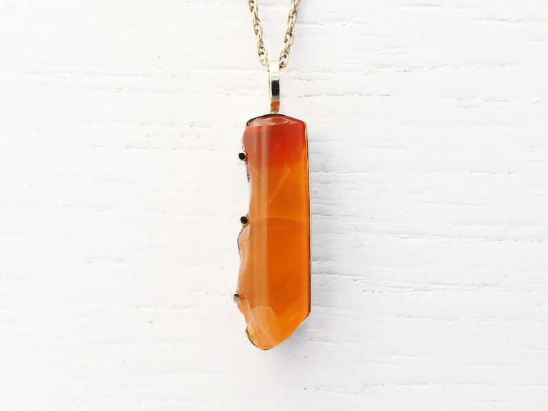 May include: A gold chain necklace with a pendant made of a large, orange carnelian gemstone. The carnelian is faceted and has a natural, irregular shape.