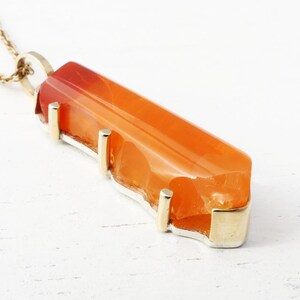 May include: A gold necklace with a large, orange carnelian pendant. The pendant is set in a gold bezel and has a rough, natural shape.