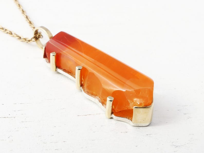 May include: A gold necklace with a large, orange carnelian pendant. The pendant is set in a gold bezel and has a rough, natural shape.