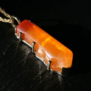 May include: A gold chain necklace with a pendant made of a long, orange carnelian stone. The stone is set in a gold frame with four prongs.