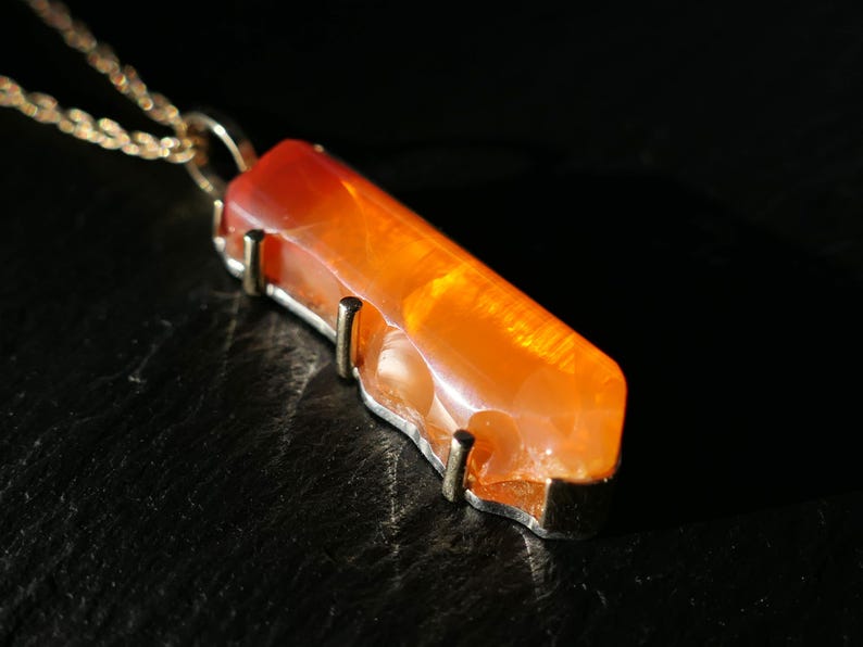 May include: A gold chain necklace with a pendant made of a long, orange carnelian stone. The stone is set in a gold frame with four prongs.