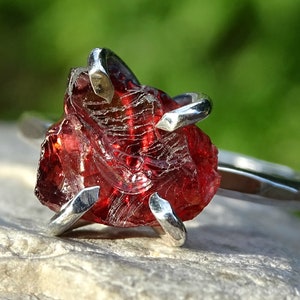 May include: A silver ring with a large, raw, red garnet gemstone. The gemstone is set in a simple, claw setting.
