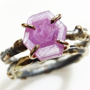 May include: A distinctive ring with a hexagonal pink gemstone, held by gold prongs. The band has a textured, organic design with silver and gold tones. The ring's design creates a striking contrast. The ring is set against a white background.