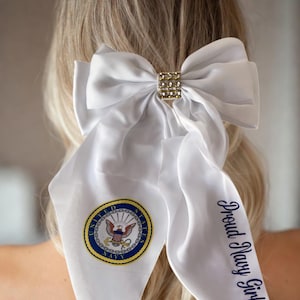 May include: White satin hair bow with a gold rhinestone center. The bow is attached to a white scarf with the US Navy emblem and the words "Proud Navy Girlfriend" in navy blue script.