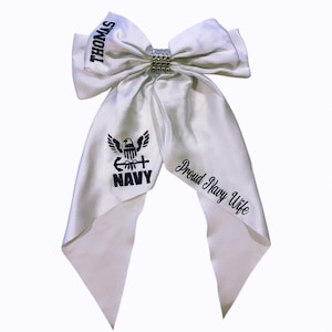 May include: A white satin bow with a rhinestone center. The bow features the name "Thomas" in black text, along with a Navy emblem and the words "Proud Navy Wife".