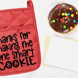 May include: Red quilted oven mitt with the text "Thanks for making me one smart cookie" in black lettering. A chocolate donut with colorful sprinkles is in the background.