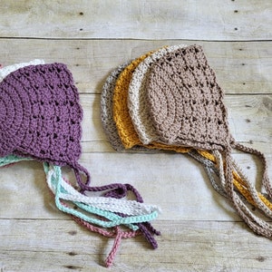 May include: A set of crocheted baby bonnets in various colors, including purple, white, mint green, and beige. The bonnets have a scalloped edge and are tied with long strings.