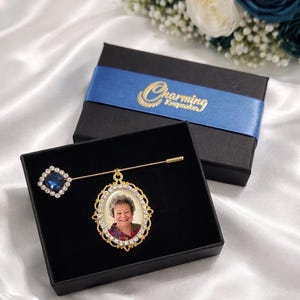 May include: A gold-toned memorial pin with a photo of a person, surrounded by a decorative frame and clear crystals. The pin features a square blue crystal accent. Presented in a black gift box with a blue ribbon that reads "Charming Keepsake".