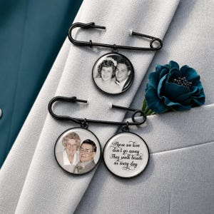 May include: Two black metal safety pins featuring circular photo charms. One pin holds a black and white portrait of a couple. The other pin holds two charms: a black and white portrait of a couple and a white disc with the black text, Those we love don't go away They walk beside us every day.