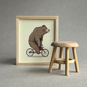 May include: A framed illustration of a brown bear riding a red bicycle. The image is displayed on a wall next to a wooden stool.
