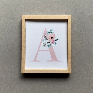 May include: A framed print with a pink letter A decorated with watercolor flowers. The frame is made of light wood.