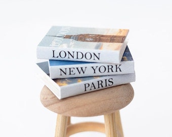 Könnte beinhalten: Ein Stapel von drei Miniaturbüchern mit den Titeln "London", "New York" und "Paris" steht auf einem kleinen Holzhocker.