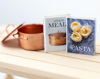 May include: Two miniature cookbooks on a wooden table. The book on the left is titled "simple MEAL" and features a copper pot on the cover. The book on the right is titled "PASTA" and features a close-up of pasta noodles.
