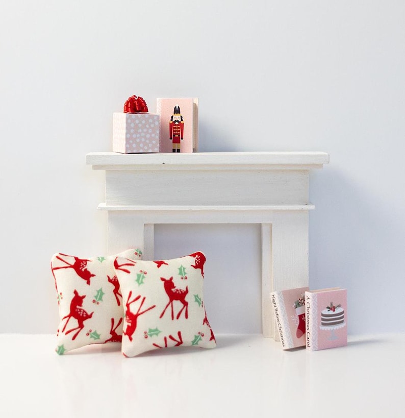 May include: Two red and white throw pillows with a reindeer and holly pattern, a white mantel with two wrapped gifts and a nutcracker figurine, and three miniature books with Christmas themes.