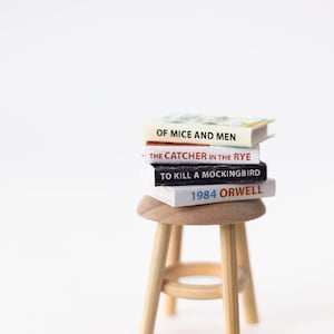 May include: A stack of four miniature books on a wooden stool. The books are titled "Of Mice and Men", "The Catcher in the Rye", "To Kill a Mockingbird", and "1984 Orwell".