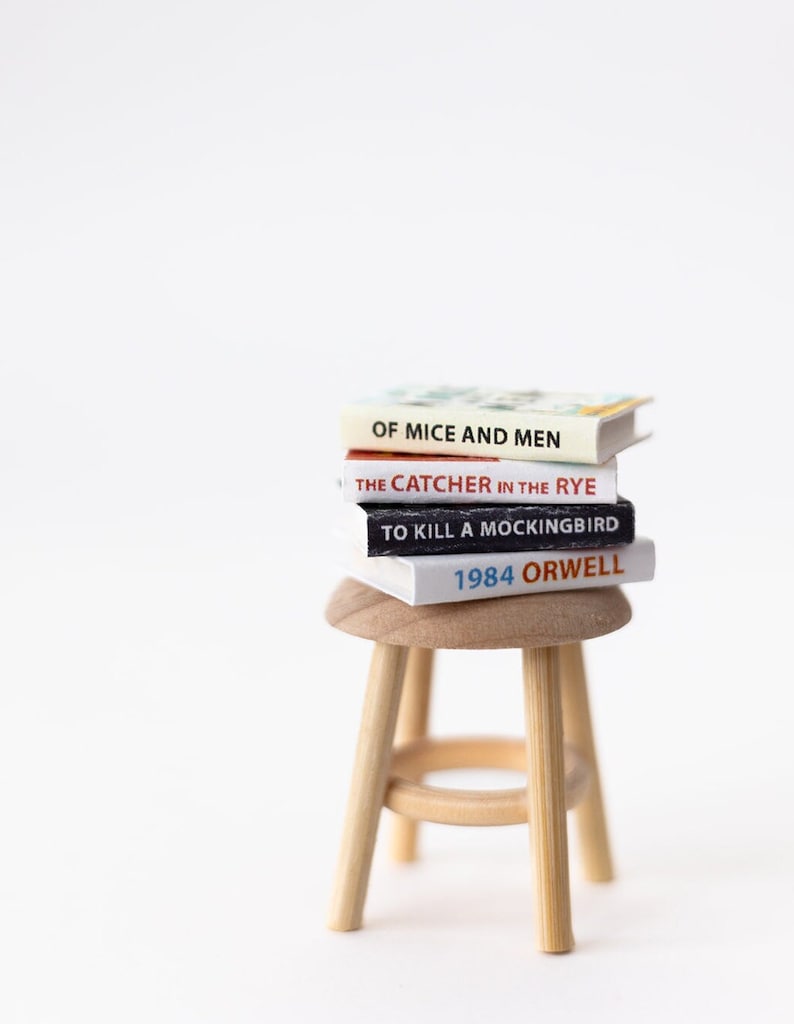May include: A stack of four miniature books on a wooden stool. The books are titled "Of Mice and Men", "The Catcher in the Rye", "To Kill a Mockingbird", and "1984 Orwell".