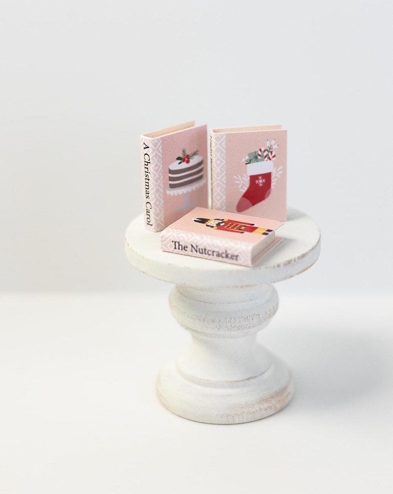 May include: Three miniature books with Christmas themes on a white pedestal table. The books are titled "A Christmas Carol", "The Nutcracker", and a book with a Christmas stocking illustration.