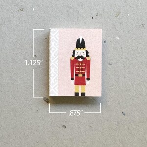 May include: A pink and white notepad with a nutcracker illustration. The nutcracker is wearing a red and gold uniform with a black hat and a white plume. The notepad is 1.125 inches tall and .875 inches wide.