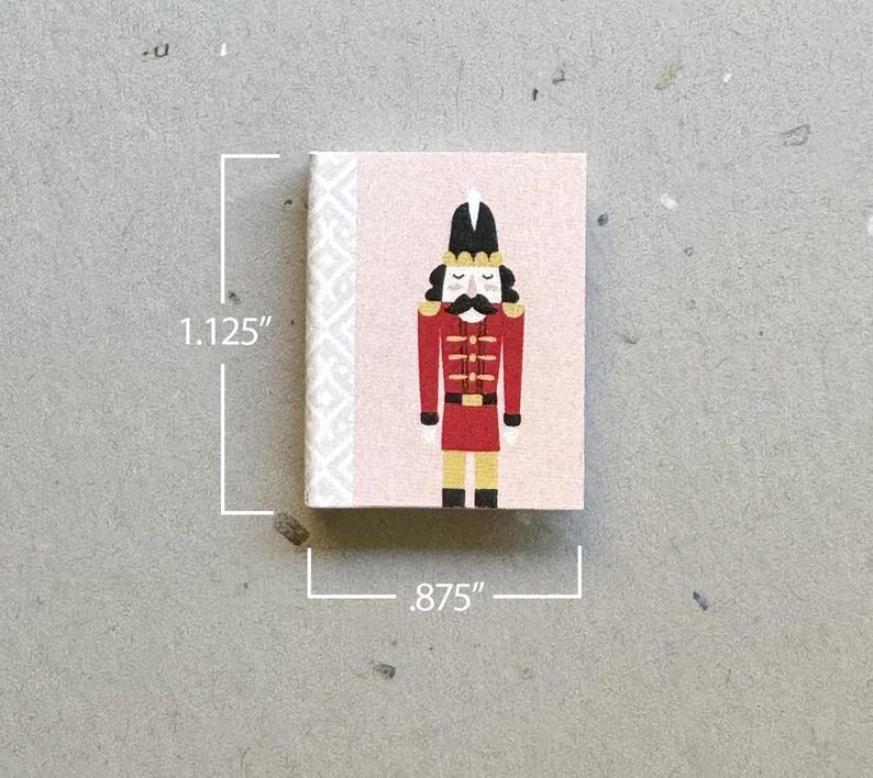 May include: A pink and white notepad with a nutcracker illustration. The nutcracker is wearing a red and gold uniform with a black hat and a white plume. The notepad is 1.125 inches tall and .875 inches wide.