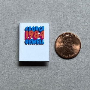 May include: A miniature book with a white cover and the title "GEORGE 1984 ORWELL" in colorful, 3D letters. The book is next to a US penny for scale.