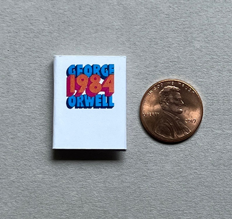 May include: A miniature book with a white cover and the title "GEORGE 1984 ORWELL" in colorful, 3D letters. The book is next to a US penny for scale.