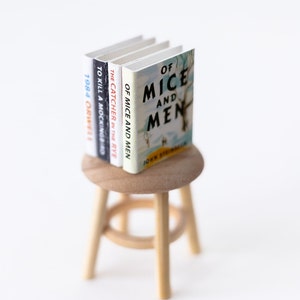 May include: A set of four classic novels, "Of Mice and Men", "The Catcher in the Rye", "To Kill a Mockingbird", and "1984", stacked on a wooden stool.
