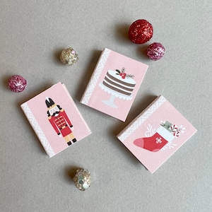 May include: Four pink gift boxes with different holiday designs. The boxes feature a nutcracker, a cake, a stocking, and a candy cane.